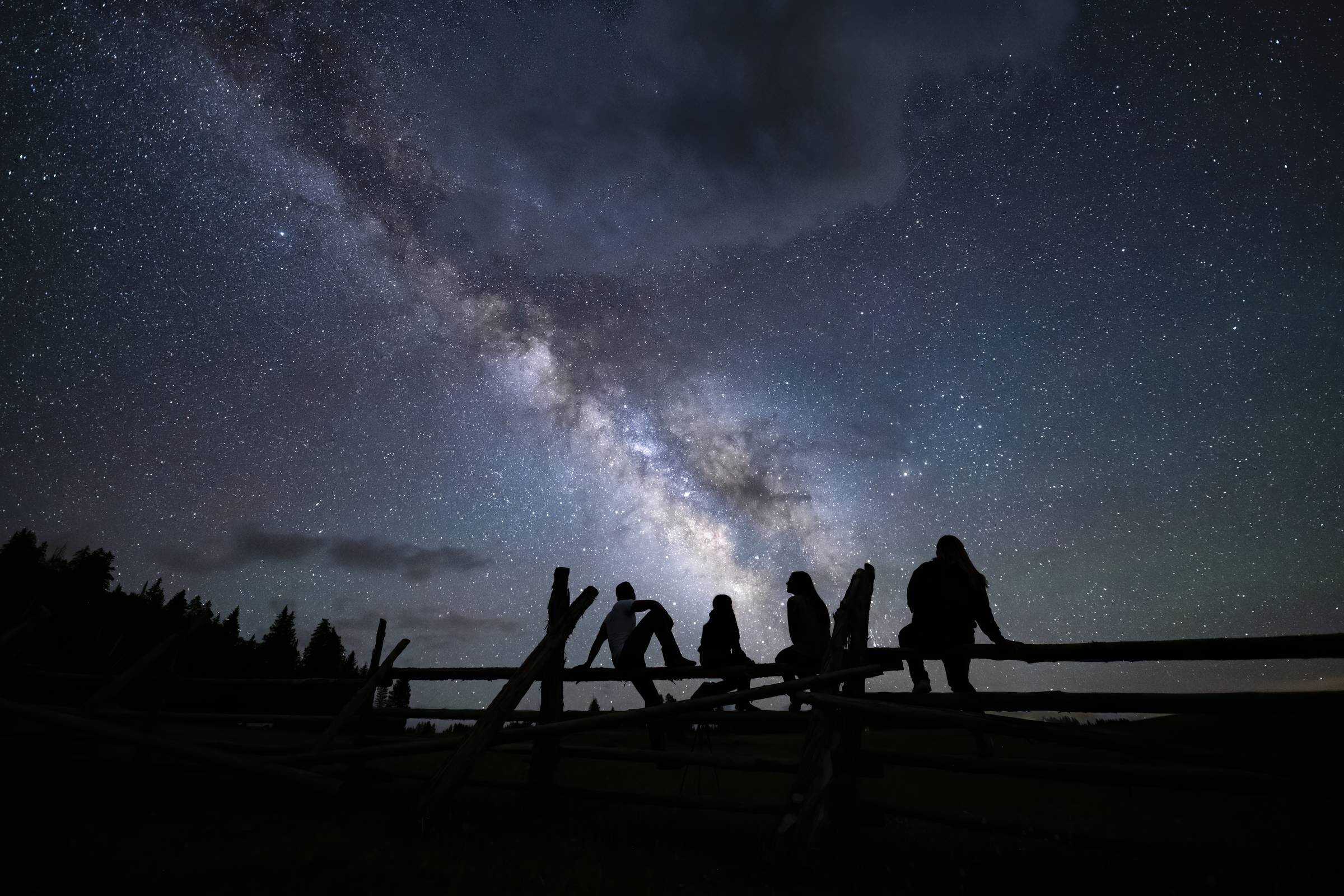 four people stargazing on a wooden fence underneath blue, purple bright milky way galaxy